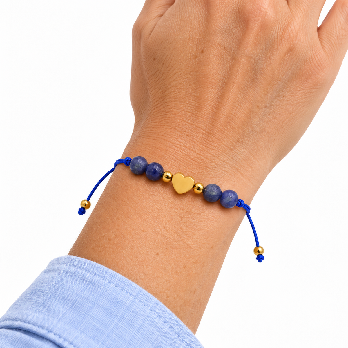 Bracelet cœur doré en sodalite porté au poignet, main relevée sur fond blanc