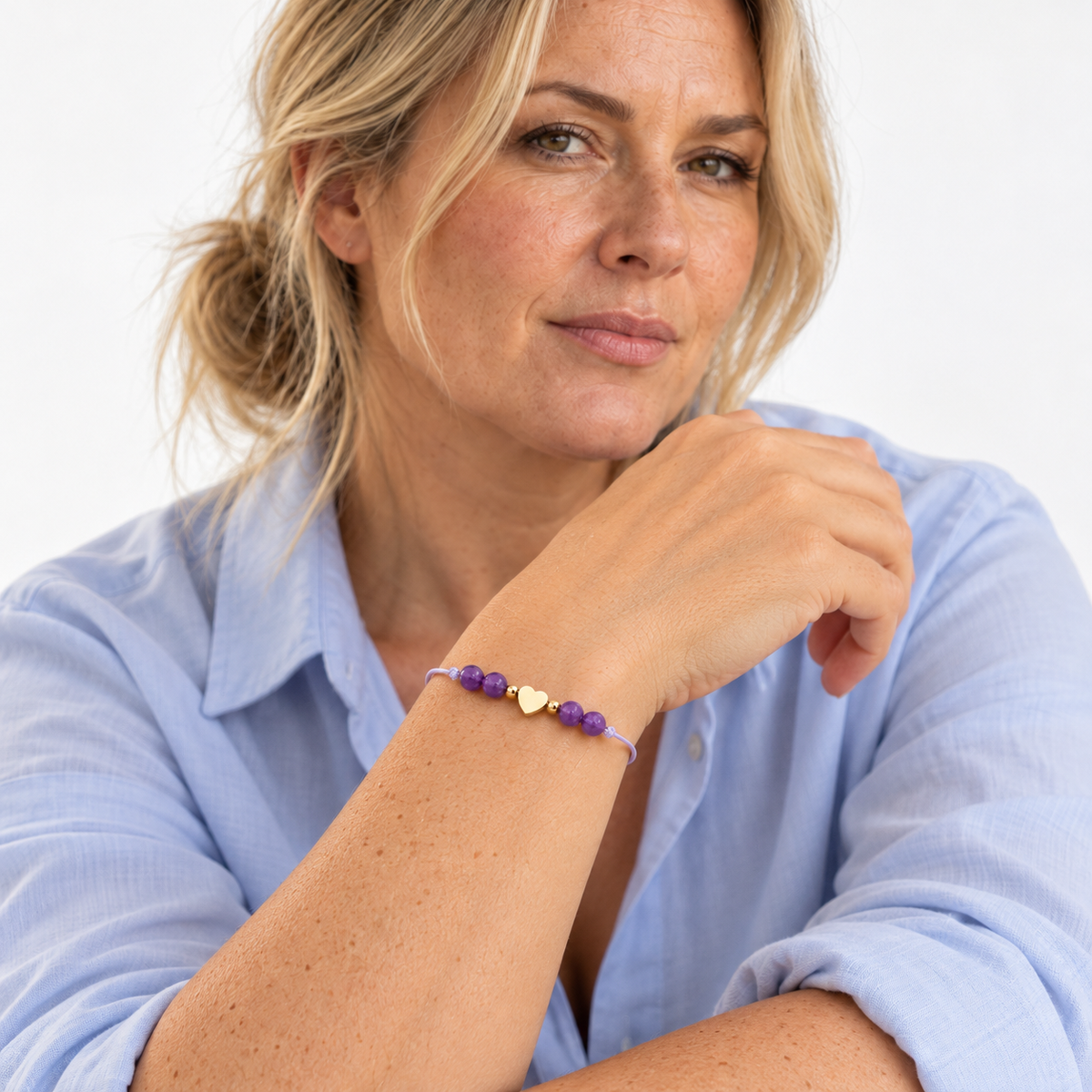 Bracelet cœur doré et améthyste porté avec une chemise bleu clair, poignet au premier plan